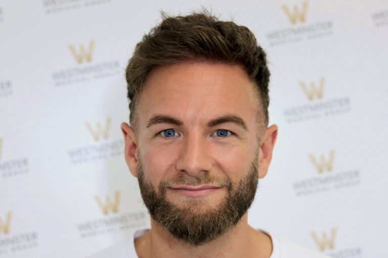 A man with a beard and short, styled hair, possibly following hair replacement therapy, smiling at the camera, standing in front of a white background with a subtle logo pattern.