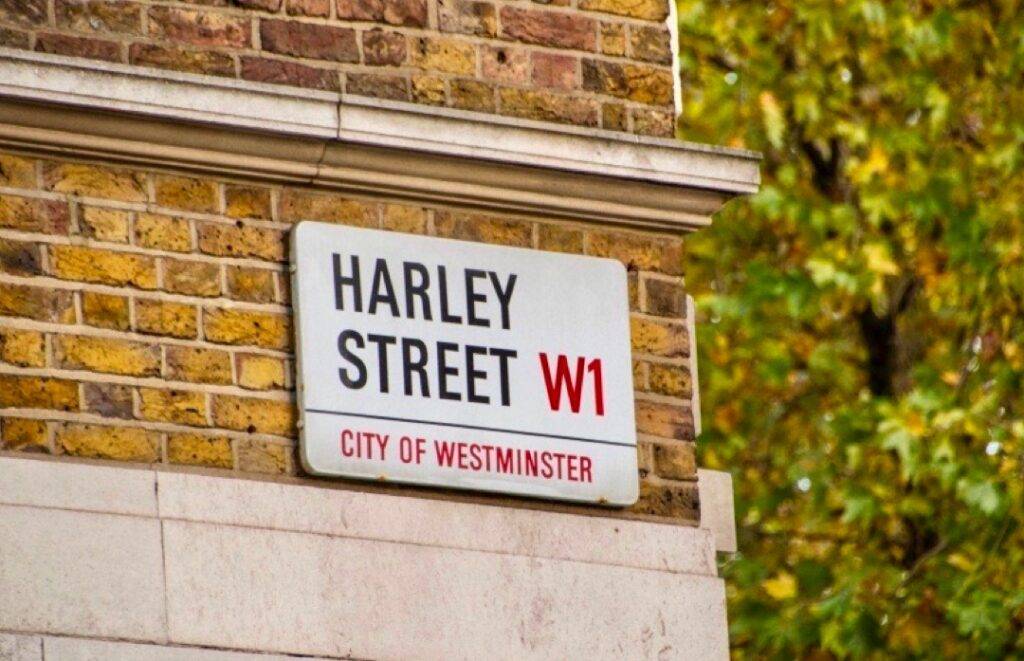 A street sign reading "harley street w1, city of westminster" mounted on a brick wall, with blurred green foliage in the background. Known for specialists in hair replacement surgery.